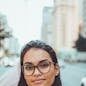 A confident woman wearing glasses smiles subtly while standing on an urban street with blurred background.